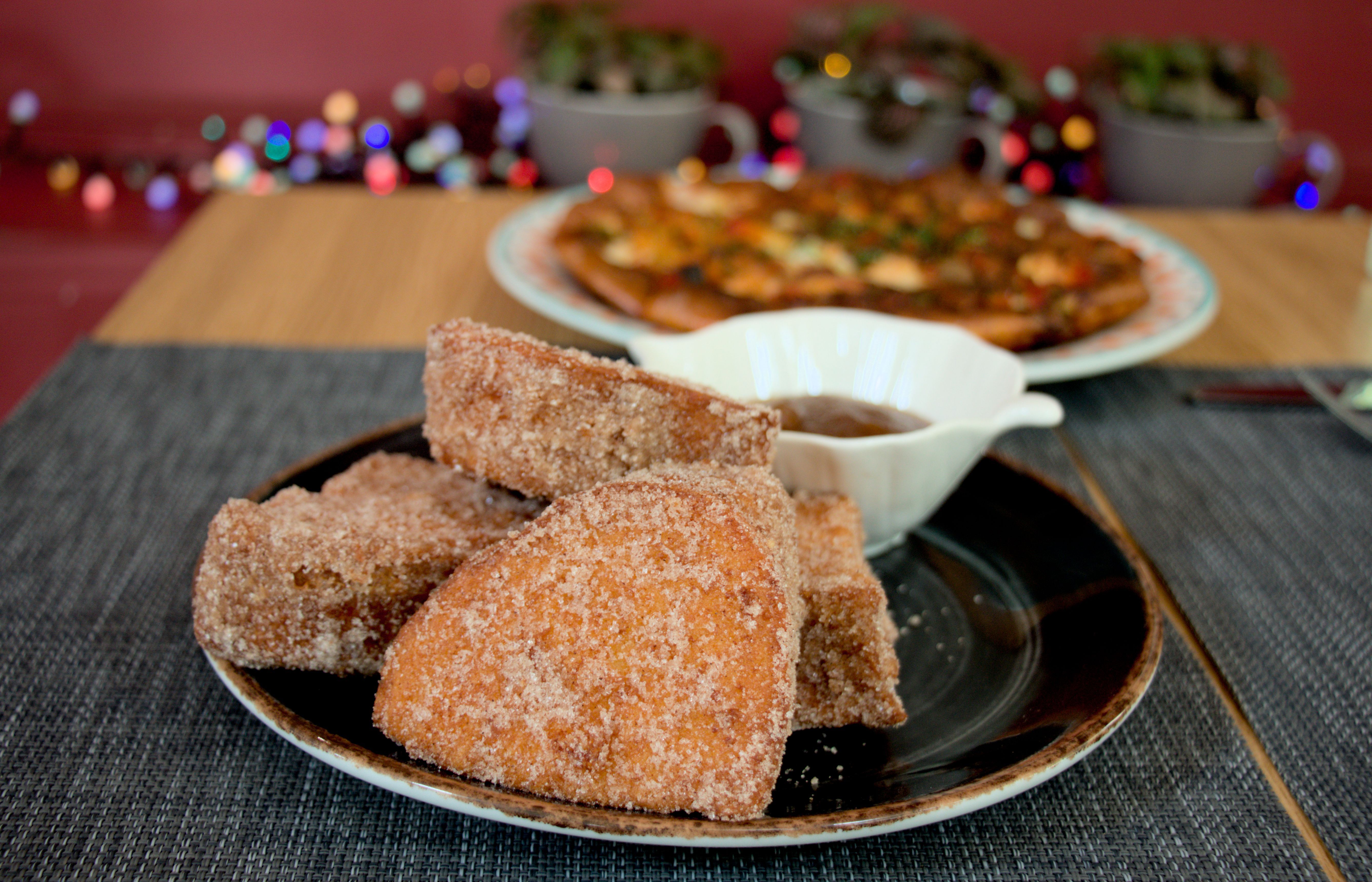 Close up, eye level view of a plate with some sugared, fried dough. In the background, you can see an out of focus pizza