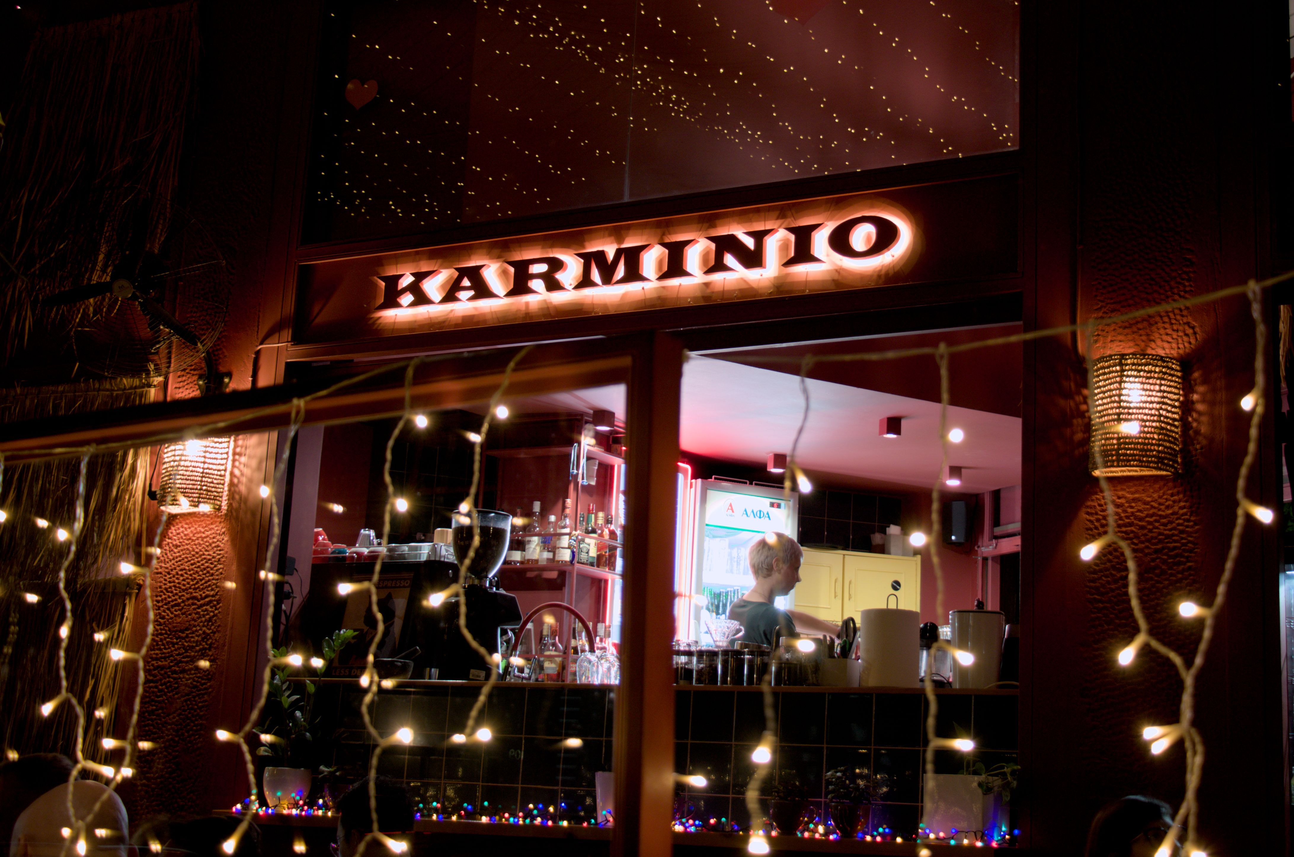 Night time photo of a bar. A prominent lit sign reads KARMINIO, over a window in which a woman is working the bar. In the foreground there are small string lights, mostly out of focus and blurred